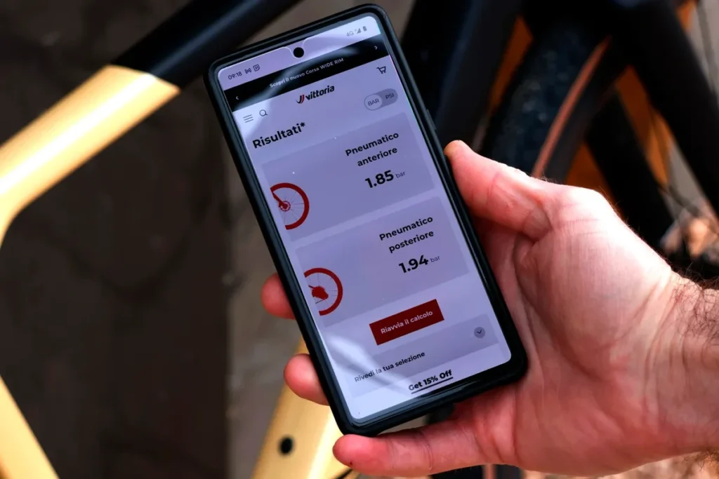 Cyclist checking bike tire pressure calculator app on smartphone beside a road bike wheel.